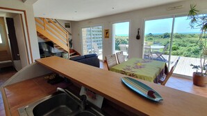 Un'ampia zona giorno con tavolo da pranzo in legno, cucina componibile con lavello, una scala e un'ampia porta scorrevole in vetro che dà su un balcone con vista sul verde e sul mare.