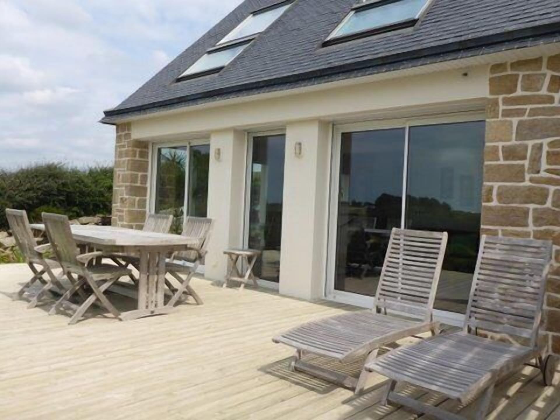 Une maison avec une façade en pierre, une terrasse en bois, une table avec des chaises et deux fauteuils de jardin.