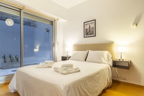 A hotel room with a bed, two bedside lamps, a framed picture of New York, and a sliding glass door leading to a balcony with plants.