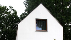 Une maison moderne aux murs blancs, dotée d’une grande porte-fenêtre coulissante, d’un petit jardin avec un arbre et d’une terrasse en bois.