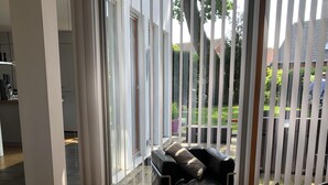 Un salon moderne avec un canapé en cuir noir, un plancher en bois et une vue sur l’extérieur à travers de grandes stores verticaux.