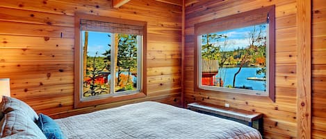 A wooden cabin bedroom with a large bed, two windows offering views of a lake and trees, and a bench by the windows.
