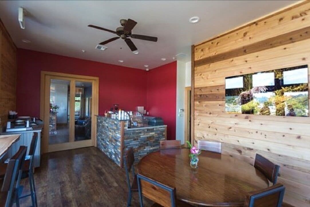 A dining area with a round wooden table and chairs, a ceiling fan, and a stone wall with a glass door leading to a kitchen.