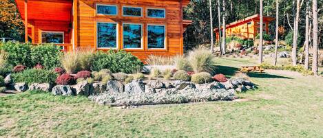 A modern wooden cabin with large windows, surrounded by a well-maintained garden and a forested backdrop.