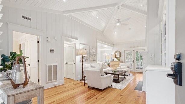 A spacious living room with hardwood flooring, white walls, and a ceiling fan. There is a white sofa, a coffee table, and a lamp.