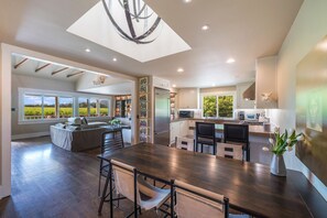 Una cocina amplia con una isla grande, un comedor con mesa de madera y vistas a un viñedo a través de ventanas panorámicas.