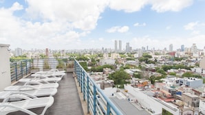 Terraço no telhado com cadeiras brancas e guarda-corpo azul, com vista para um horizonte urbano com edifícios e áreas verdes.