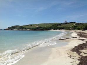 A beach with clear water, a sandy shore, and a hill with vegetation and a lighthouse in the distance.