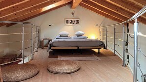 Une chambre à coucher aménagée dans un espace de type « loft », avec un plafond haut, des poutres en bois, un lit avec des oreillers, un tapis tissé et un petit banc.