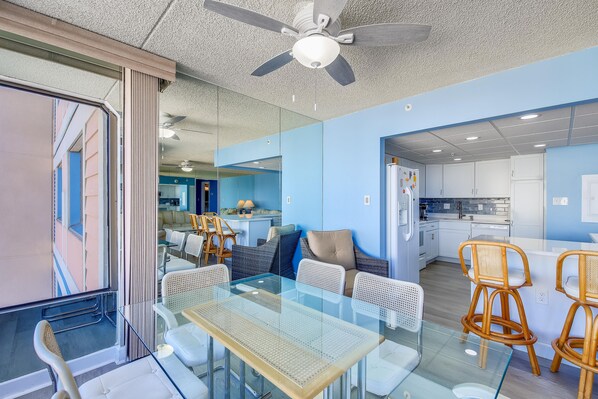 Un interior moderno con una mesa de comedor de vidrio, sillas de mimbre, un ventilador de techo y una zona de cocina visible a través de una puerta abierta.