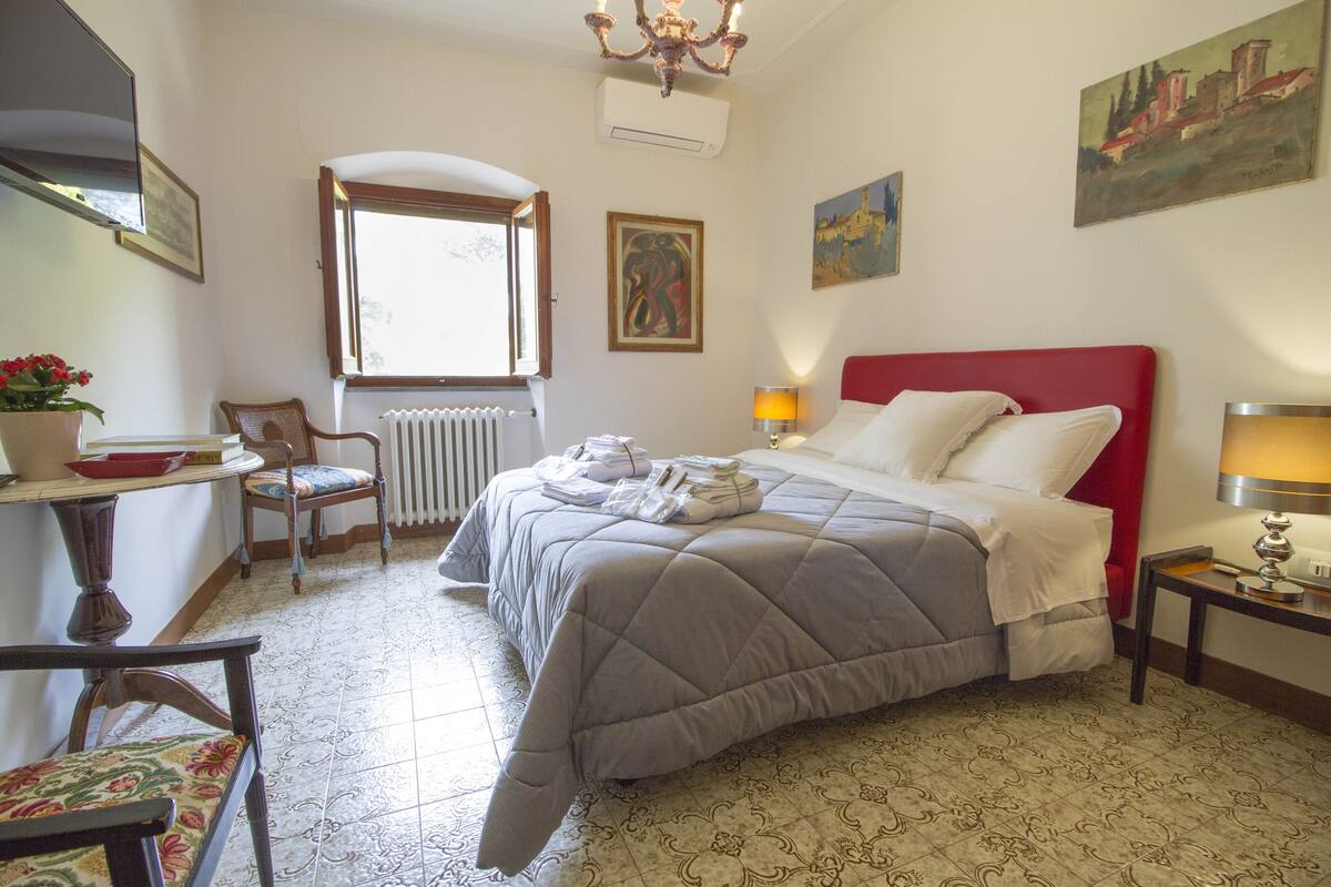 A bedroom with a bed, a chair, a table with a lamp, and a window.