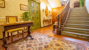 A traditional interior with a wooden staircase, a green door, a bench, a table with a vase, and a patterned rug.