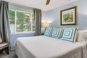Une chambre à coucher avec un lit, une table de chevet avec une lampe, une fenêtre avec des rideaux et un tableau encadré au mur.