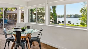 A dining area with a glass table and chairs, large windows with a view of a lake and outdoor seating.