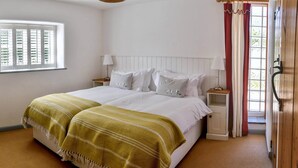 A bedroom with a bed, two bedside tables, a window with blinds, a door with a curtain, and a wooden floor. - Superb detached Grade II listed barn conversion with private hot tub, close to coast (Ottery St Mary)