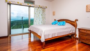 Une chambre à coucher avec un lit en bois, une table de chevet, une commode et une grande porte-fenêtre coulissante donnant sur un balcon avec une vue panoramique.