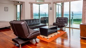 Un salon spacieux avec un canapé en cuir, un fauteuil noir et une table basse en verre. De grandes fenêtres offrent une vue sur la mer et les collines.