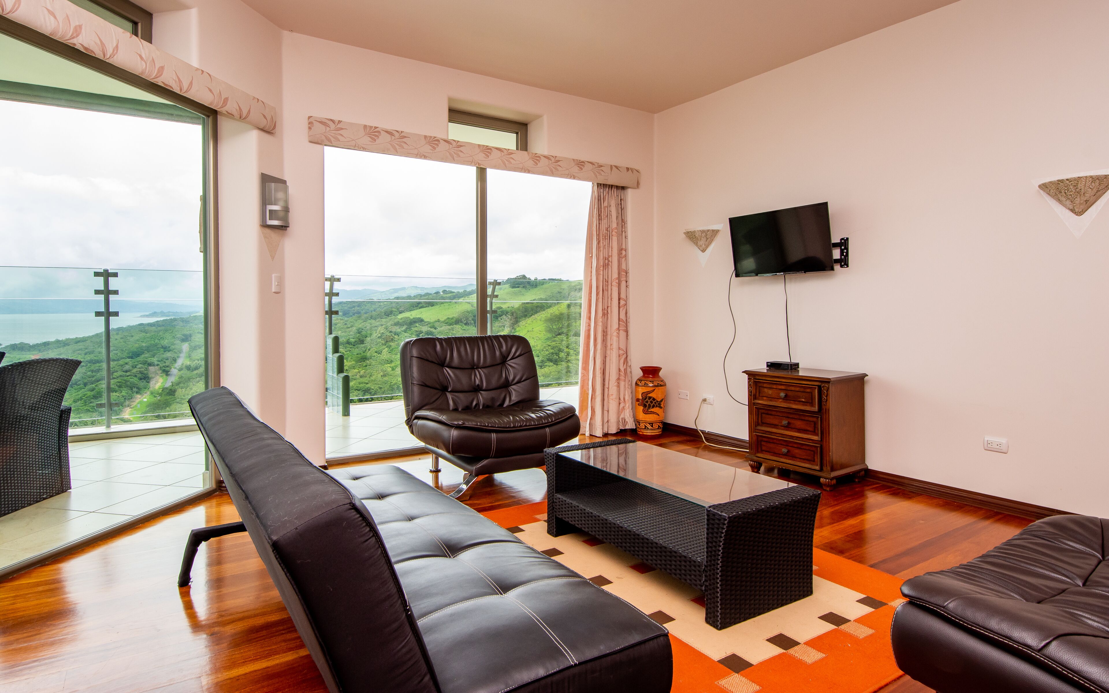 Amplia sala de estar con un ventanal que ofrece vistas panorámicas, un sofá de cuero, una mesa de centro y un televisor de pantalla plana montado en la pared.
