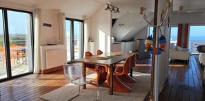 Un espace repas comprenant une table, des chaises et une vue sur la mer, grâce à de grandes fenêtres.