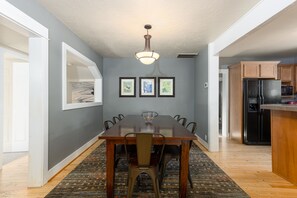 Un coin repas avec une table en bois, des chaises et un lustre. Des tableaux encadrés ornent le mur, et on aperçoit une cuisine au fond.