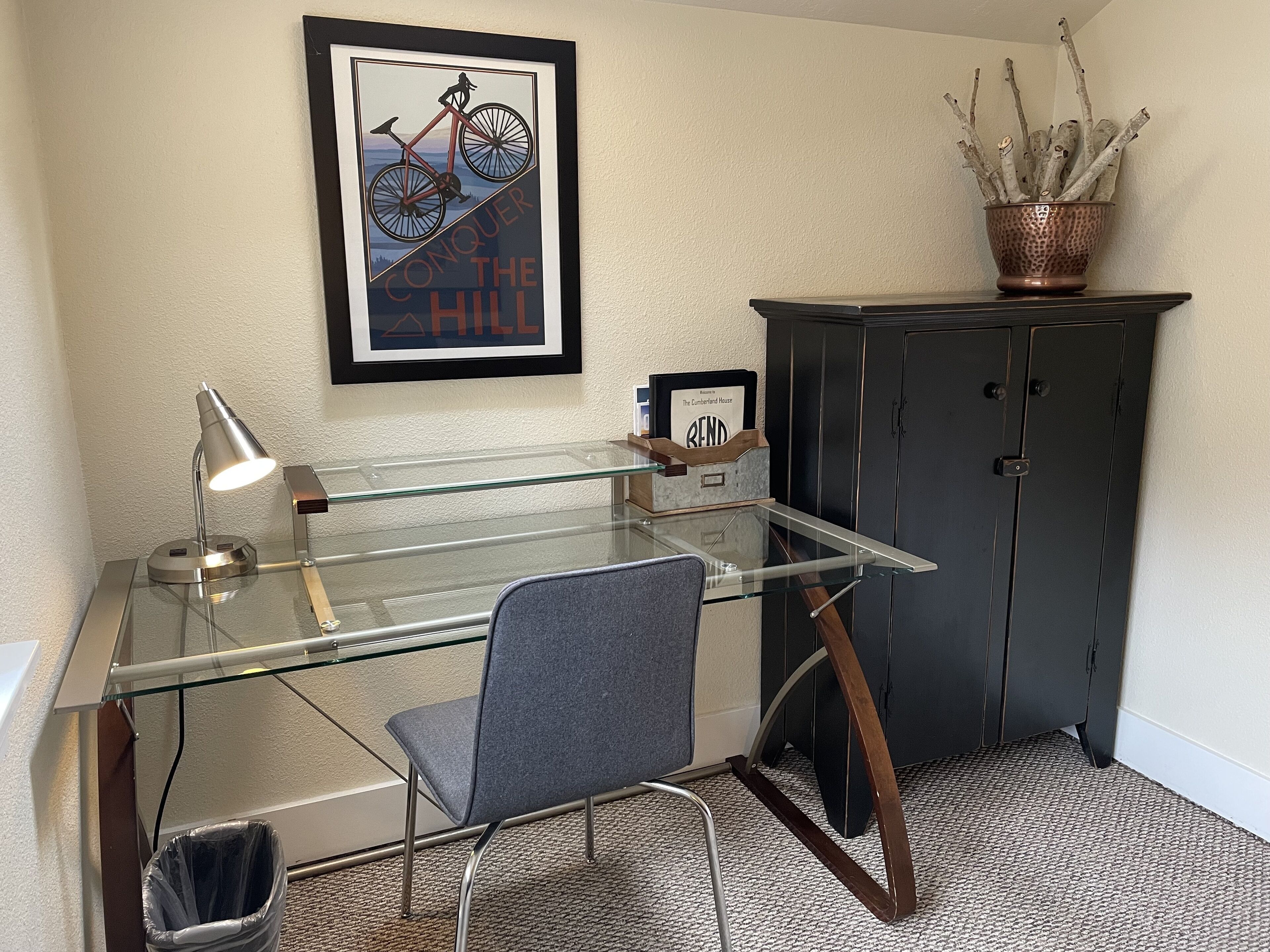 Un espace de bureau moderne avec un bureau en verre, une chaise, une affiche encadrée, un meuble et une plante en pot.