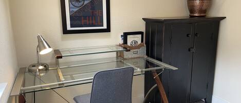 Un espace de bureau moderne avec un bureau en verre, une chaise, une affiche encadrée, un meuble et une plante en pot.