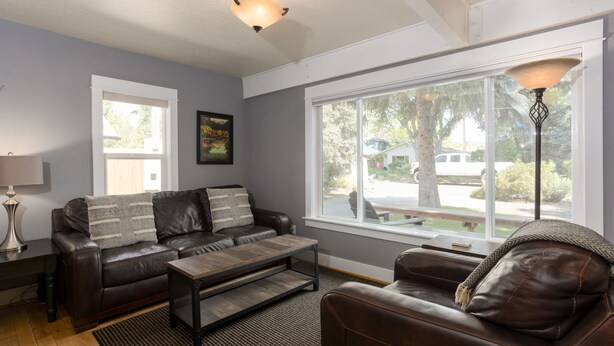 A living room with a brown leather sofa, a coffee table, and a lamp. There is a window with a view of a tree and a house. - Spacious Layout │West Side │Walk Everywhere │Hot Tub │One Block to River (Bend)