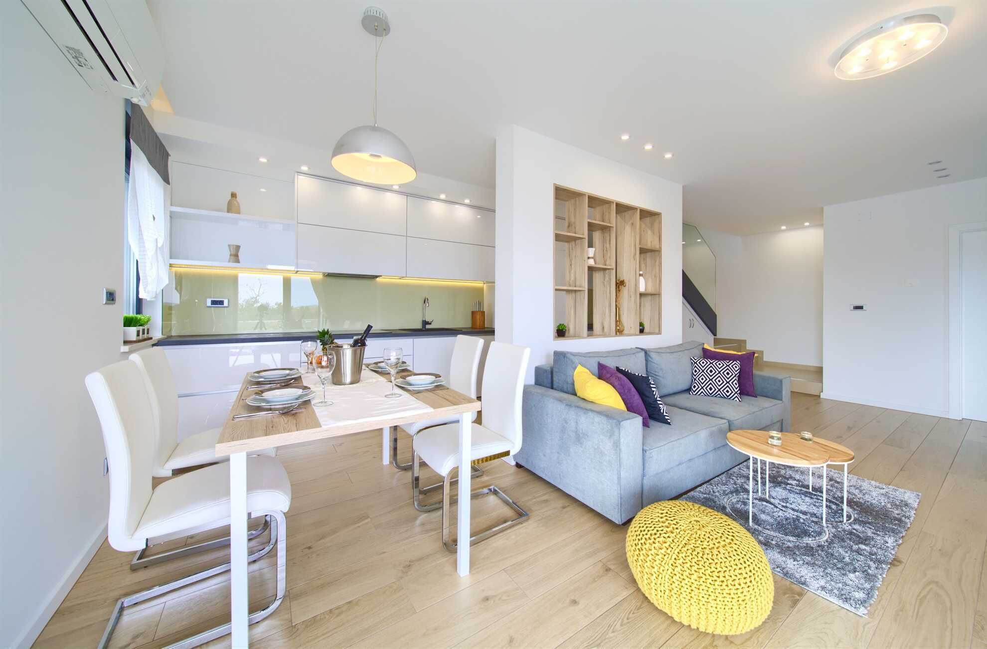 Ein modernes Wohnzimmer mit einem grauen Sofa, weißen Stühlen, einem Holzespeisetisch für vier Personen und einem kleinen runden Couchtisch.