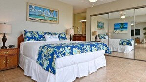 A hotel room with a bed, a nightstand, a mirror, and a painting of a beach scene. - BeachFront 2BD w Stunning Ocean View-Paki Maui 424 (Lahaina)