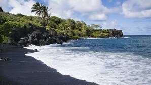 A black sand beach with waves crashing onto the shore, surrounded by lush greenery and palm trees. - BeachFront 2BD w Stunning Ocean View-Paki Maui 424 (Lahaina)