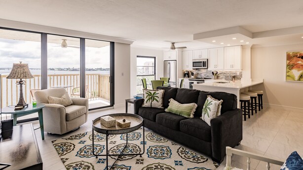 Un vaste espace de vie comprenant un canapé noir, une table basse ronde et un tapis à motifs. Un balcon avec vue sur la ville est visible à travers de grandes fenêtres.