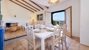 A dining area with a white table set for four, wooden chairs, a hanging light fixture, and a view through an open door to the outdoors.