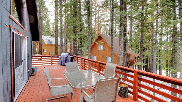 A wooden deck with a table and chairs, surrounded by a red railing, overlooking a forested area. - Christmas Cabin (Yosemite National Park)