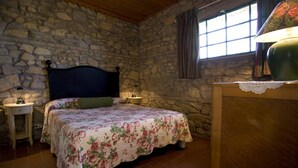Ein Schlafzimmer mit Steinsandsteinwand, einem Bett mit floralem Überwurf, einem Nachttisch mit Lampe und einem Fenster mit Vorhängen.