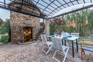 Um pátio externo com piso de pedra, forno de pedra, cadeiras brancas listradas e uma mesa posta para refeições.