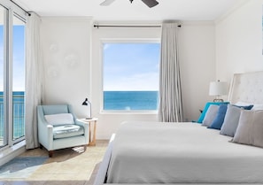 A bedroom with a large bed, a chair, a nightstand, a lamp, and a view of the ocean.