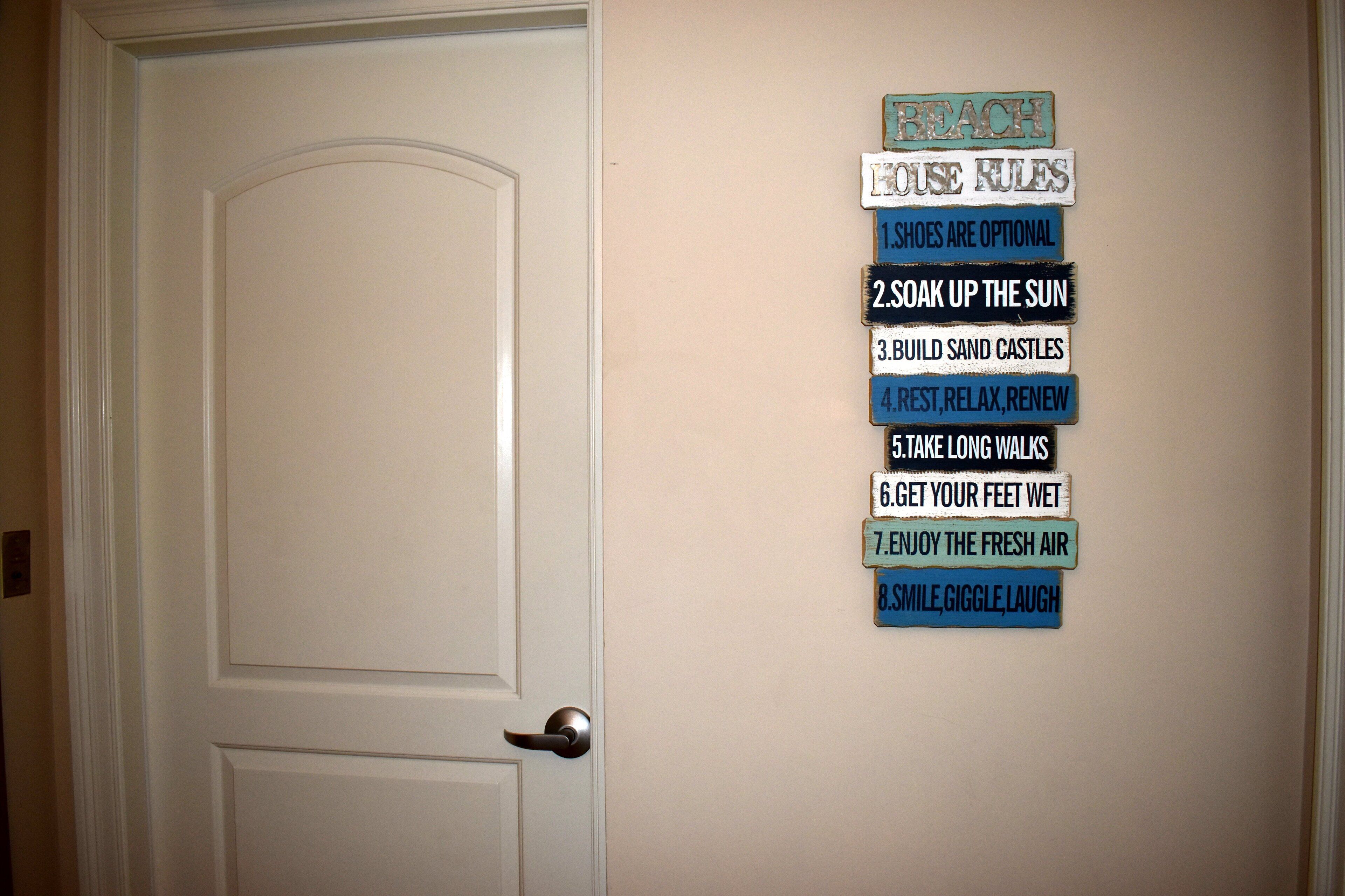 Une porte blanche avec une poignée en métal et un mur orné de huit panneaux bleus et blancs indiquant les règles à respecter dans la maison de vacances.