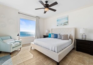 Un dormitorio con una cama grande, un ventilador de techo, una ventana con vista al océano, una silla y una mesita de noche con una lámpara.