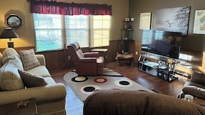 A living room with a sofa, armchair, and a coffee table. There is a TV on a stand and a window with blinds. - Fish cleaning station, screened porch, covered front porch, fire pit, 3 BR    (Hornbeak)