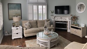 Een woonkamer met een hoge plafond, een open haard, een televisie en een groot raam met luiken.
