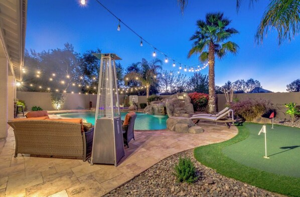 Un jardin avec une piscine, des chaises longues, une pergola illuminée de guirlandes lumineuses et un petit terrain d’entraînement au putting.