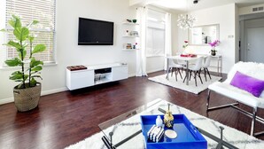 A modern living room with a flat-screen TV, a dining area with a white table and chairs, a large potted plant, and a blue tray on a glass coffee table. - ★Sunny Condo 5min from the Beach★ (Laguna Niguel)