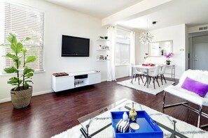 A modern living room with a flat-screen TV, a dining area with a white table and chairs, a large potted plant, and a blue tray on a glass coffee table.