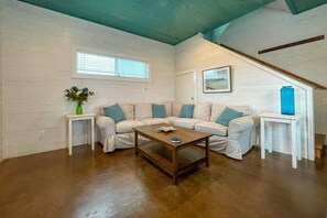 Ein Wohnzimmer mit einem beigefarbenen Ecksofa, einem Holztisch, einem kleinen Beistelltisch mit einer blauen Vase, einem Fenster mit Jalousien und einem gerahmten Bild an der Wand.