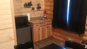 A compact kitchen with a microwave, sink, and cabinets. There is a table and chairs in the dining area. - Cabin 5 (Garden City)
