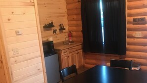 A wooden cabin interior with a dining table, chairs, a kitchen area, and a 'WELCOME' sign on the wall. - Cabin 5 (Garden City)