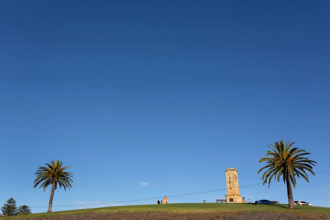 A tower on a grassy hill with palm trees, clear sky, and a few people in the distance.