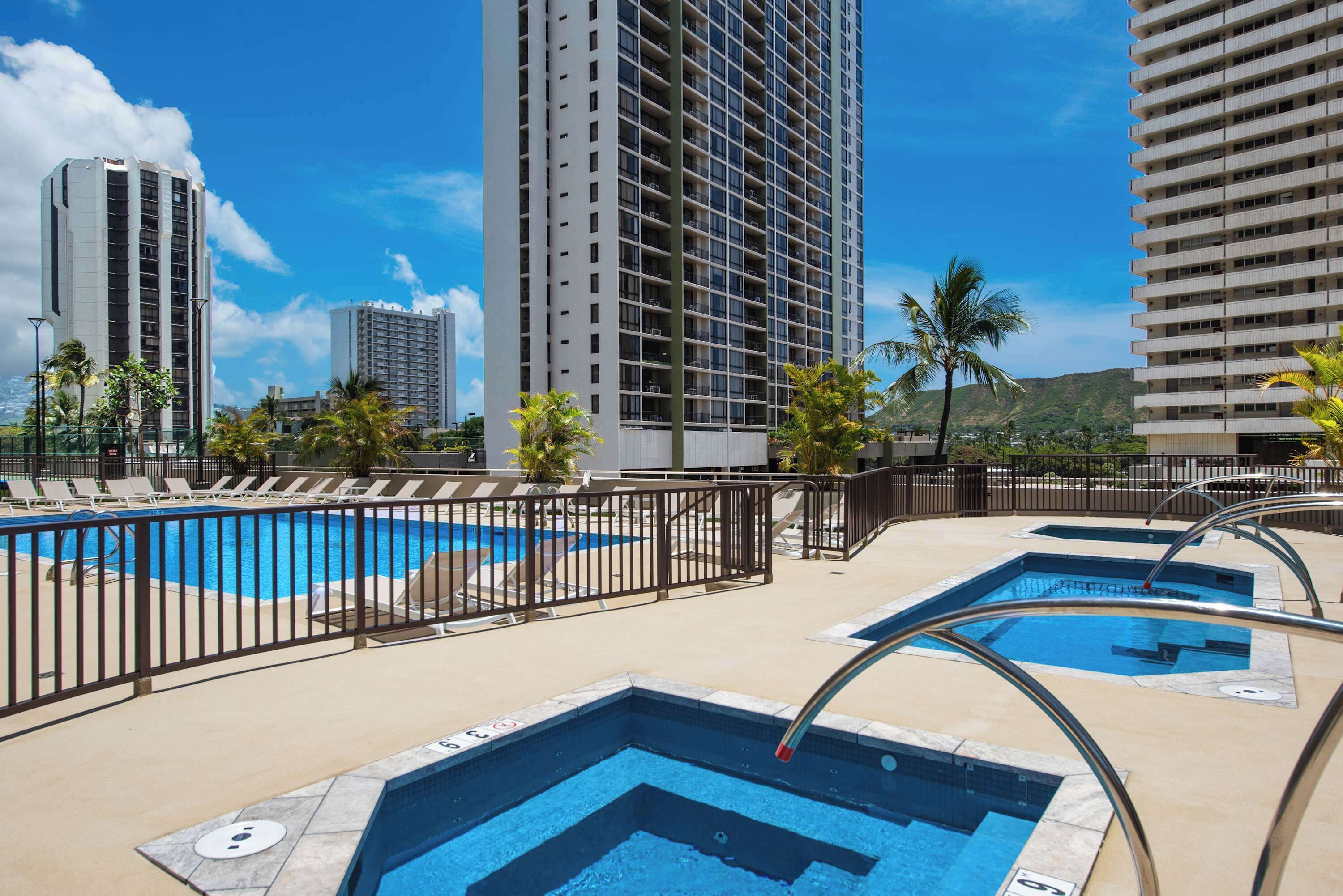 A hotel pool area with multiple pools, a fence, and tall buildings in the background.