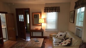 A living room with a wooden coffee table, a beige sofa with animal-themed cushions, a patterned rug, a small side table with a lamp, and a window with blinds.