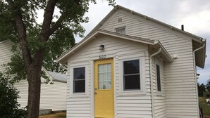A small, single-story house with a yellow door and white siding, numbered 507, featuring a front porch and a small window.
